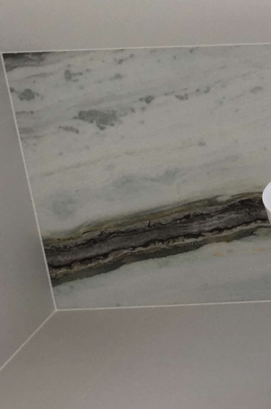 Fragment ściany wykończonej płytami imitującymi marmur z wyraźnym, ciemniejszym pasmem. Widoczny narożnik i fragment sufitu. Minimalistyczny design łazienki.
