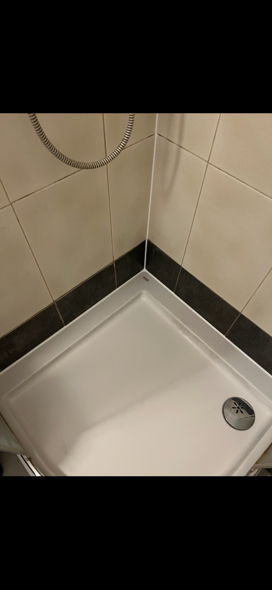 Biały brodzik kabiny prysznicowej z odpływem, obłożony beżowymi płytkami z czarnym pasem. W rogu widoczny przewód prysznicowy.