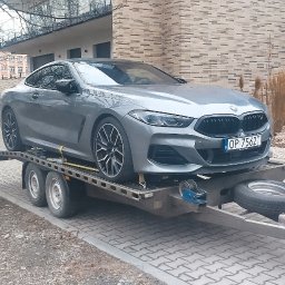 Zarasprint LTD - Szary samochód BMW na lawecie, zabezpieczony żółtymi pasami transportowymi. W tle budynek mieszkalny i drzewa bez liści. Tablica rejestracyjna widoczna.