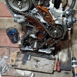 Zarasprint LTD - Silnik samochodowy z łańcuchem rozrządu na drewnianym podłożu, obok narzędzia warsztatowe: klucze, wiertarka. Widok z góry, oświetlenie warsztatowe.