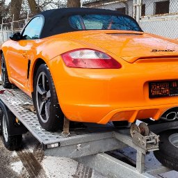 Zarasprint LTD - Pomarańczowy Porsche Boxster na lawecie, zabezpieczony pasami transportowymi. Widok z tyłu, zima w tle. Tablica rejestracyjna zasłonięta.