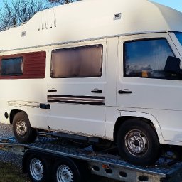 Zarasprint LTD - Biały kamper Mercedes-Benz na niskopodwoziowej lawecie, widok z boku na tle zabudowań i drzew. Transport pojazdów kempingowych.