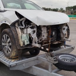 Zarasprint LTD - Uszkodzony, biały Peugeot 2008 bez przedniego zderzaka, na lawecie. Widoczne elementy silnika. W tle inne pojazdy i zieleń.