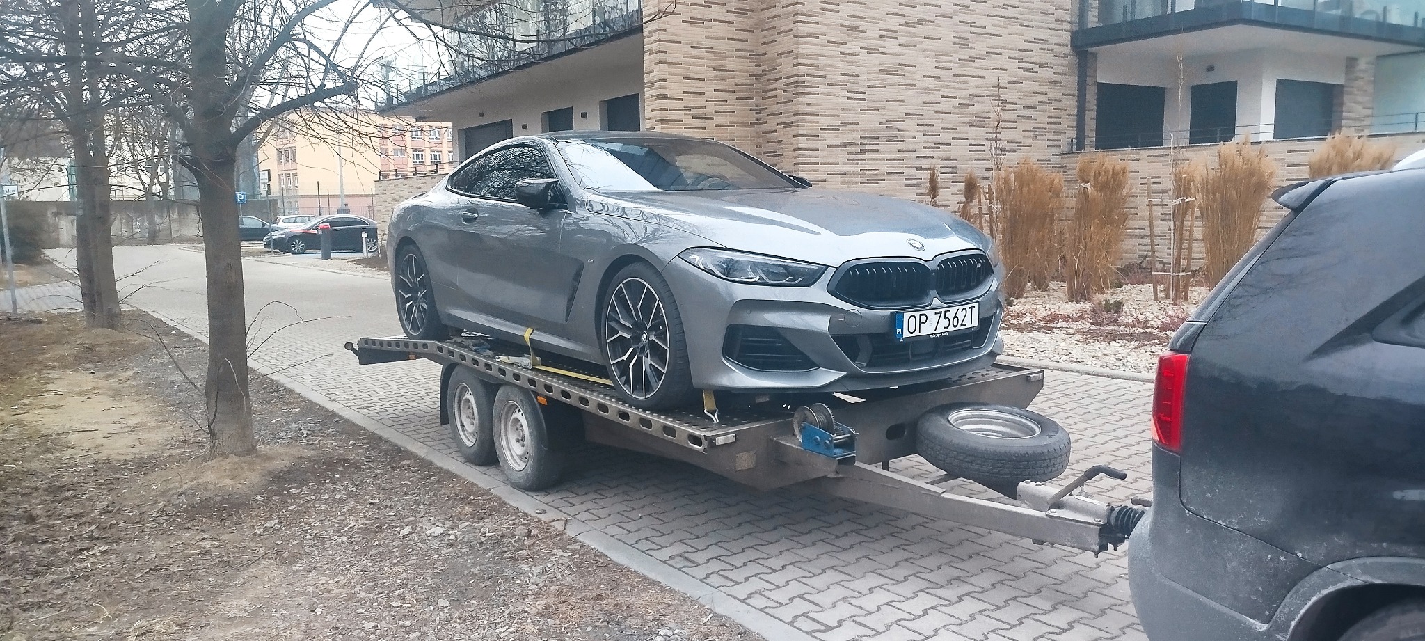 Szary samochód BMW na lawecie, zabezpieczony żółtymi pasami transportowymi. W tle budynek mieszkalny i drzewa bez liści. Tablica rejestracyjna widoczna.