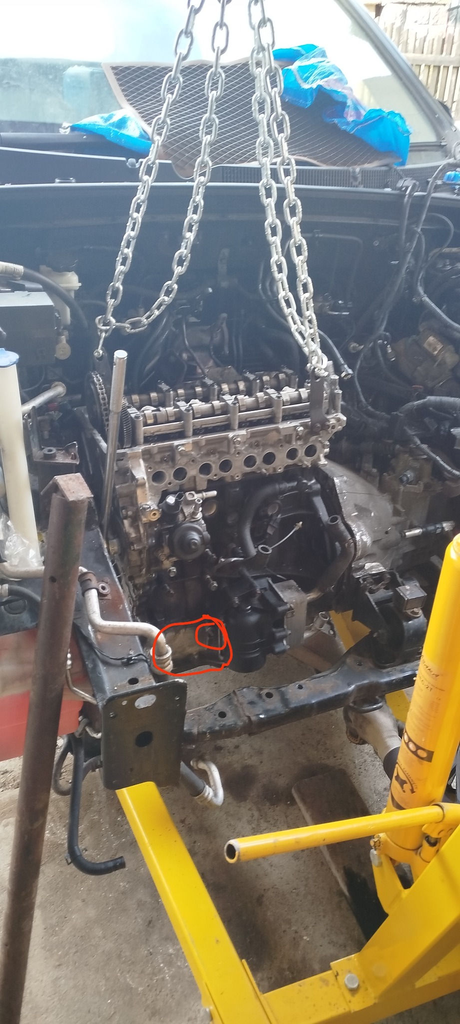 Silnik samochodowy zawieszony na łańcuchu w warsztacie, widoczne elementy konstrukcyjne pojazdu i narzędzia. Detal: czerwone okręgi na silniku.