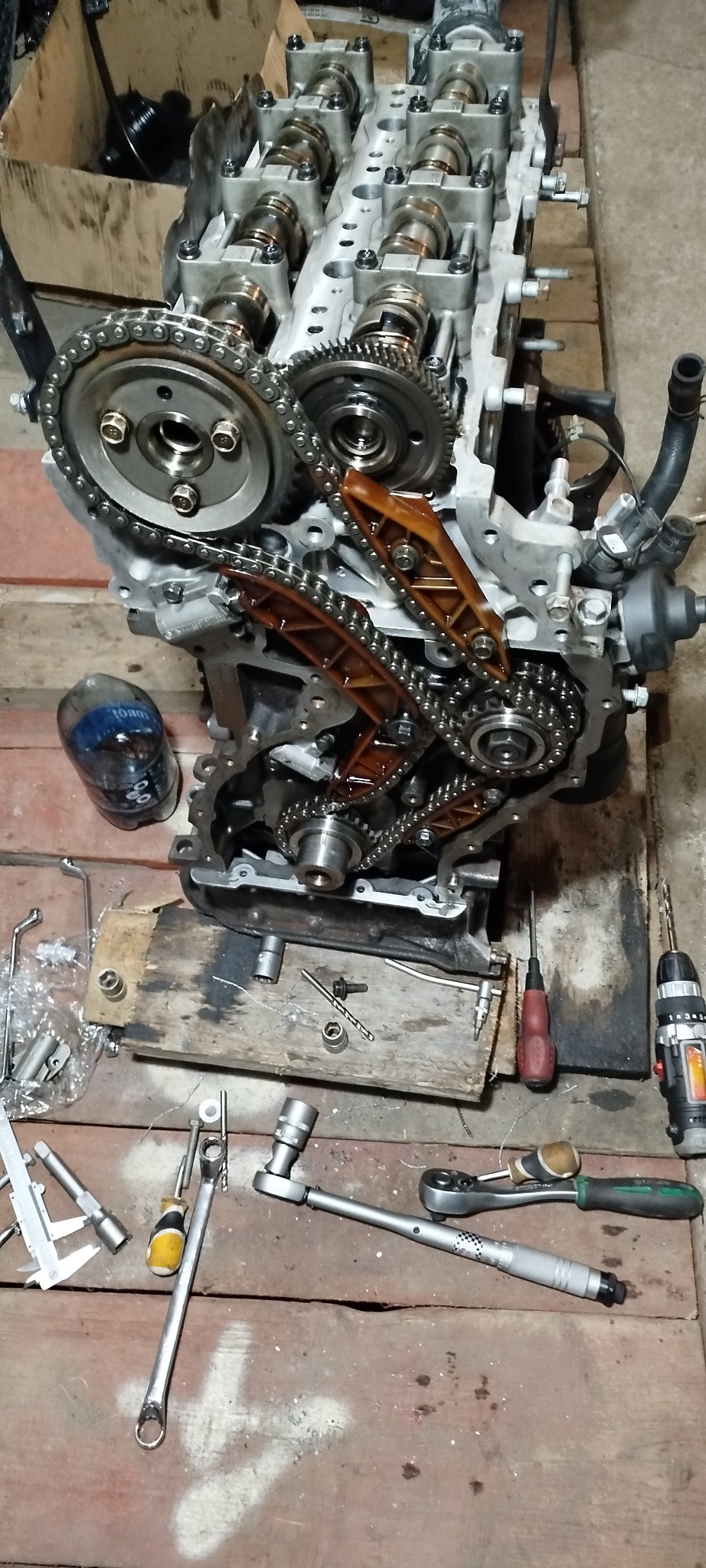 Silnik samochodowy z łańcuchem rozrządu na drewnianym podłożu, obok narzędzia warsztatowe: klucze, wiertarka. Widok z góry, oświetlenie warsztatowe.