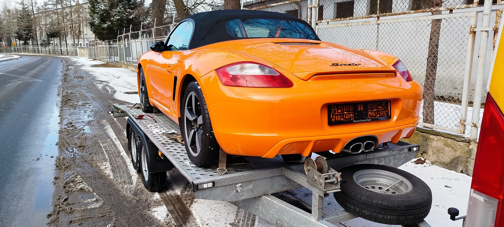 Pomarańczowy Porsche Boxster na lawecie, zabezpieczony pasami transportowymi. Widok z tyłu, zima w tle. Tablica rejestracyjna zasłonięta.