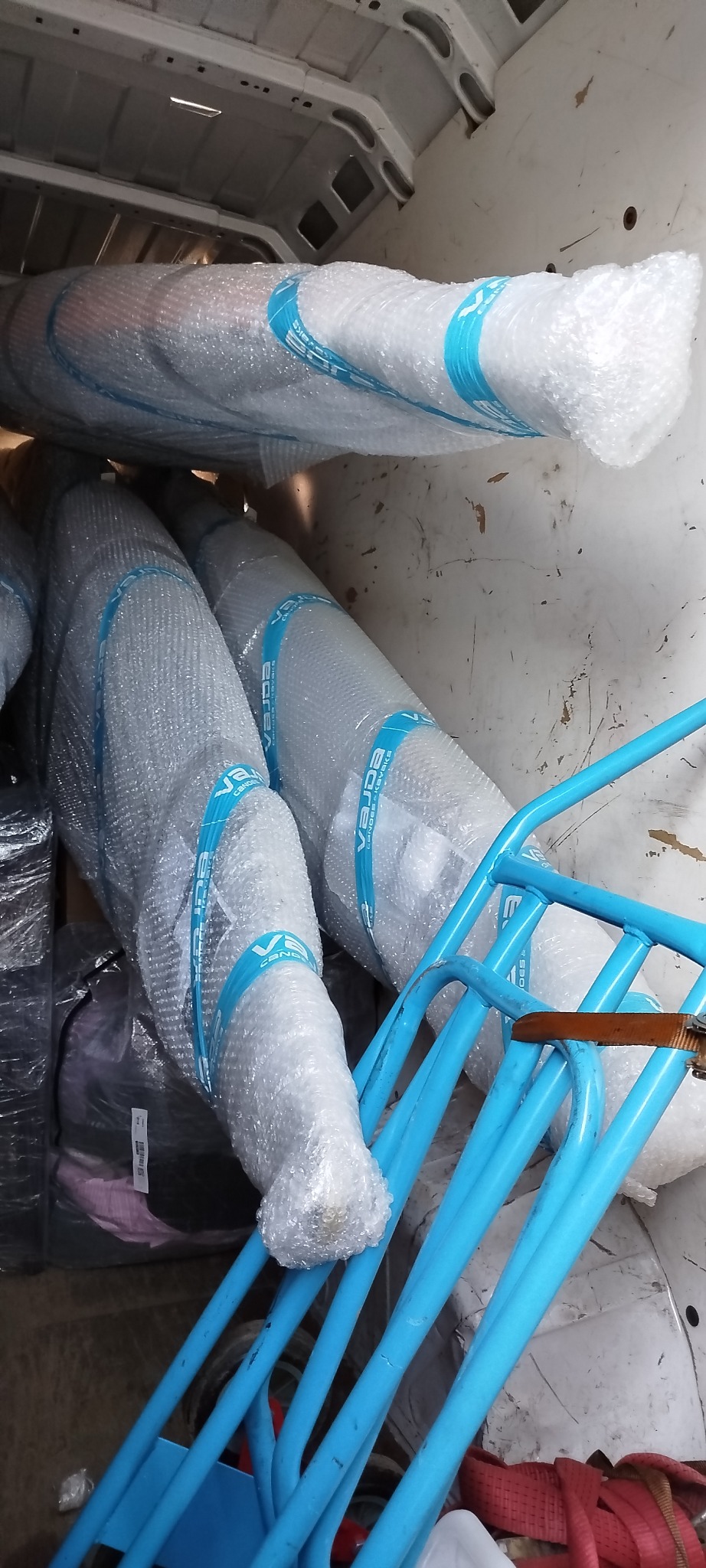 Wnętrze busa z ładunkiem: zabezpieczone folią bąbelkową rolki z niebieskimi paskami i niebieski wózek transportowy. Widok z góry, oświetlenie dzienne.