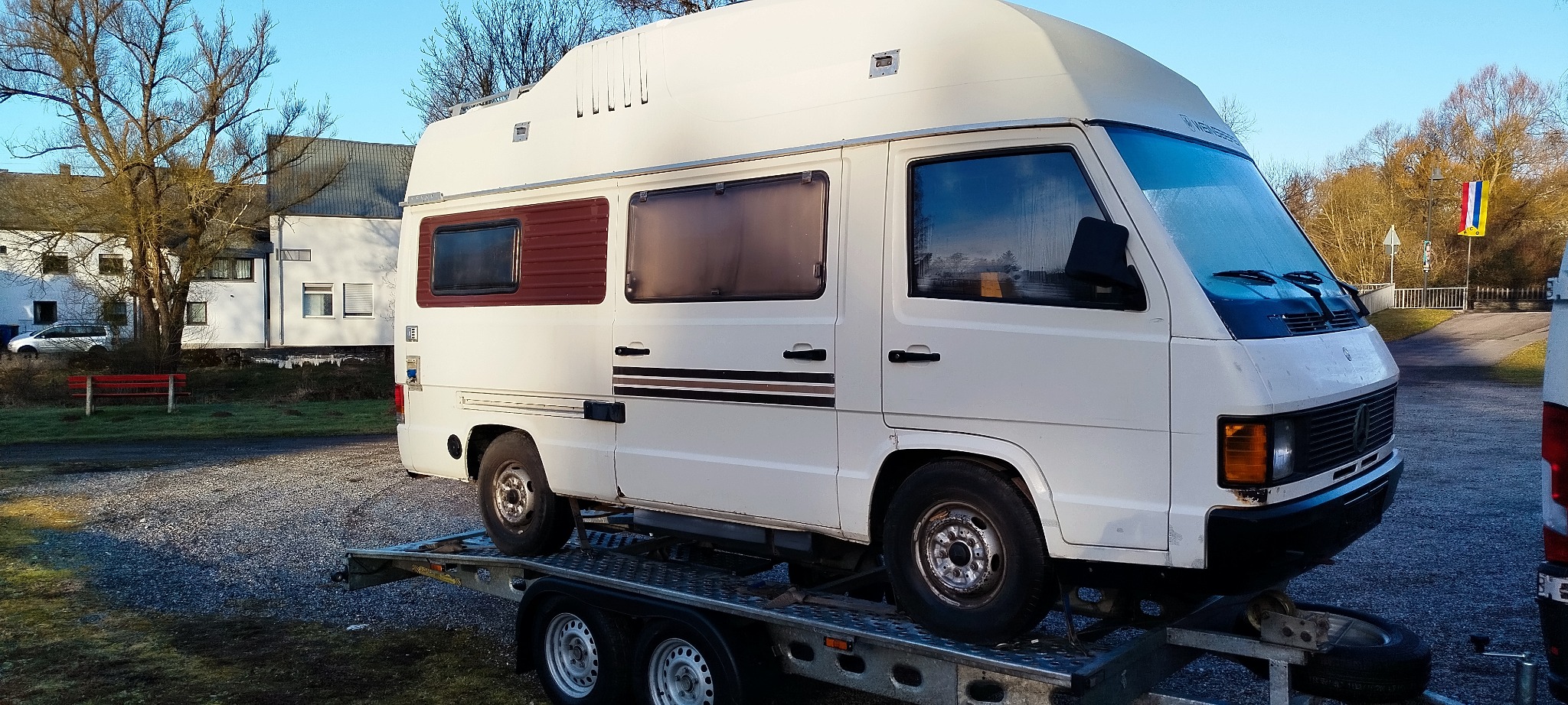 Biały kamper Mercedes-Benz na niskopodwoziowej lawecie, widok z boku na tle zabudowań i drzew. Transport pojazdów kempingowych.