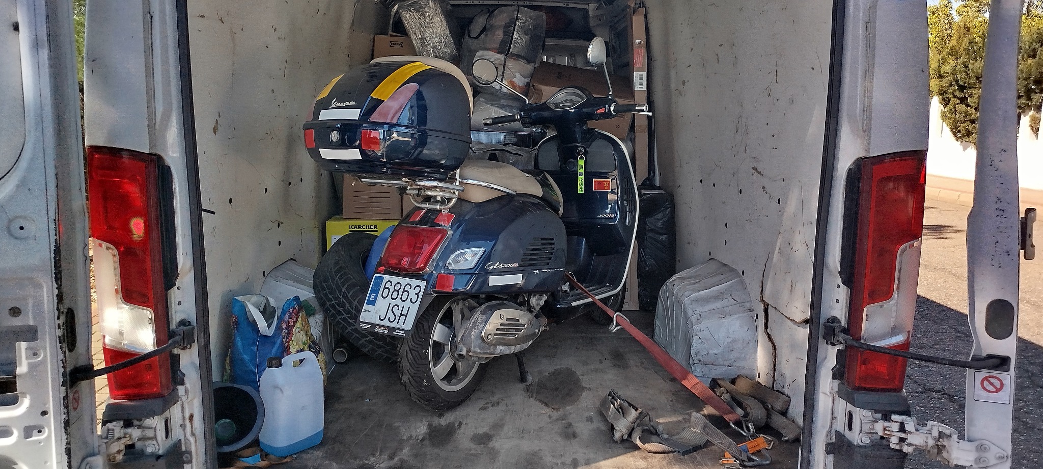 Wnętrze busa z załadowanym skuterem Vespa GTS 300, zabezpieczonym pasami transportowymi. Widoczne kartony, opona i inne przedmioty. Otwarta przestrzeń ładunkowa.