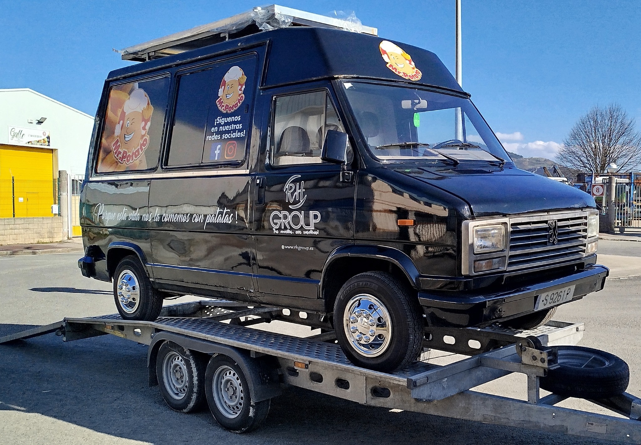 Czarna furgonetka z reklamą 'Mr. Potato' na lawecie, widok z boku. Tablica rejestracyjna widoczna. Błękitne niebo w tle. Chromowane felgi.