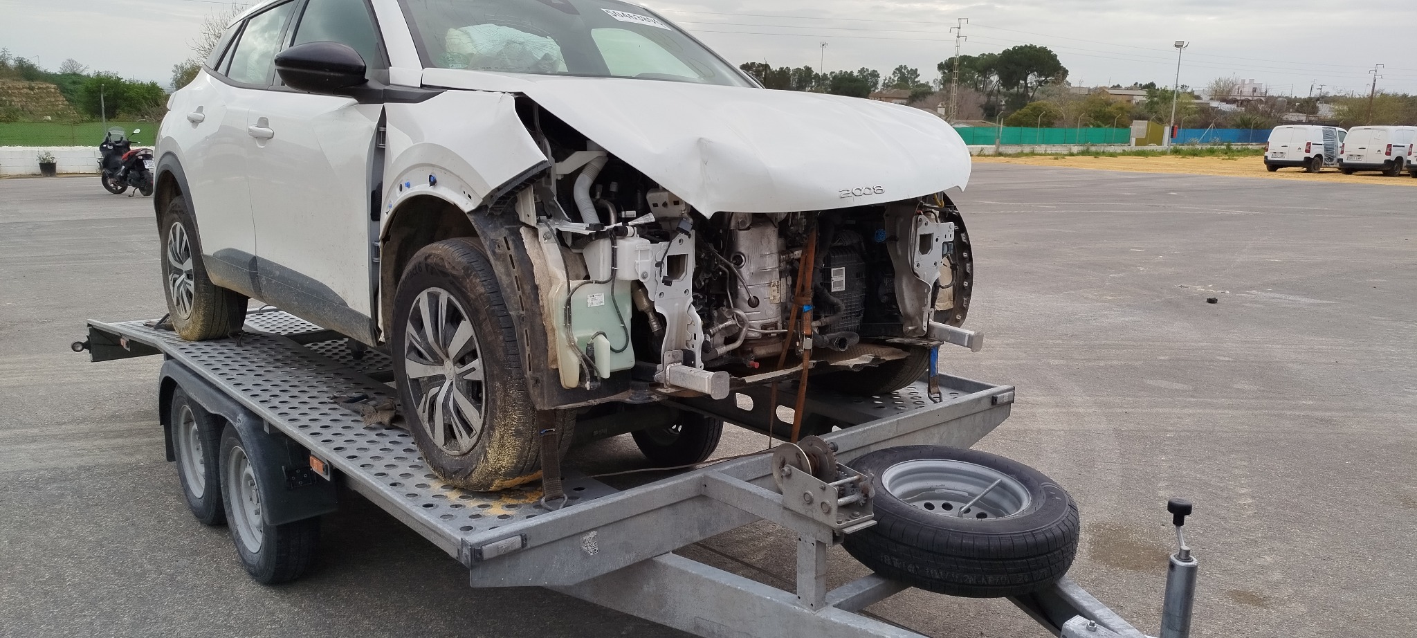 Uszkodzony, biały Peugeot 2008 bez przedniego zderzaka, na lawecie. Widoczne elementy silnika. W tle inne pojazdy i zieleń.