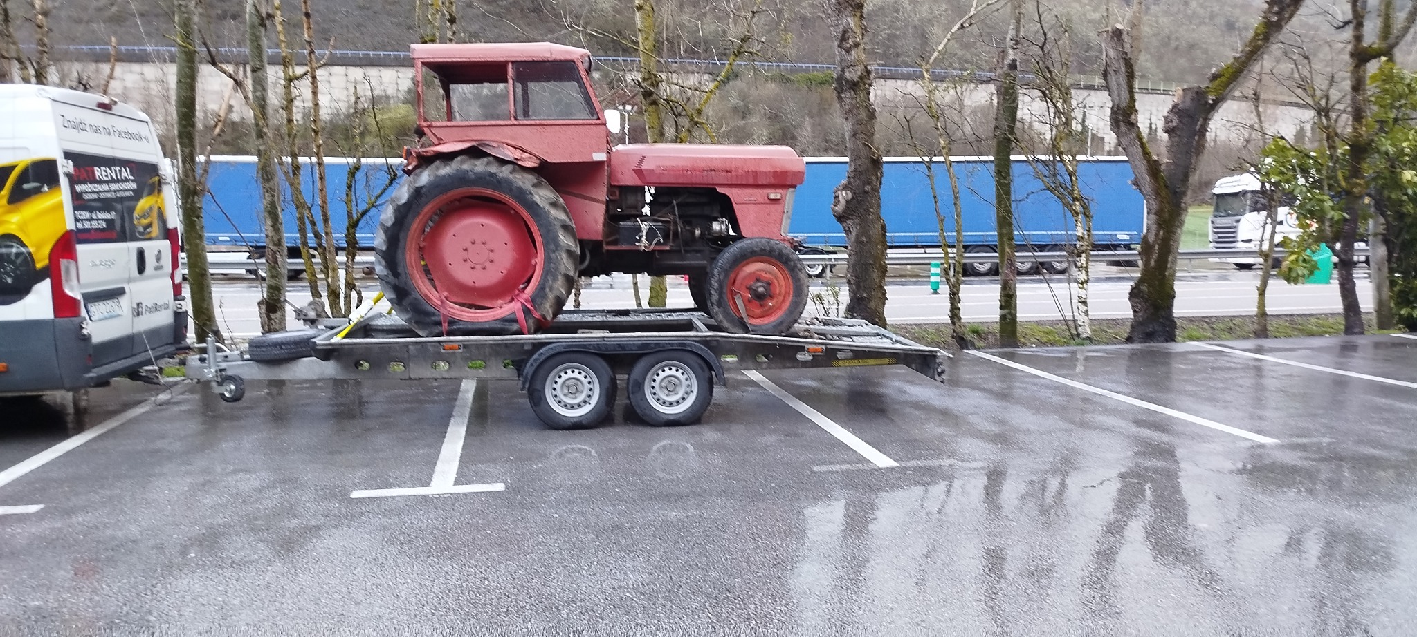 Czerwony traktor zabezpieczony pasami na niskopodwoziowej przyczepie, zaparkowanej obok busa z reklamą firmy transportowej. W tle droga i drzewa.