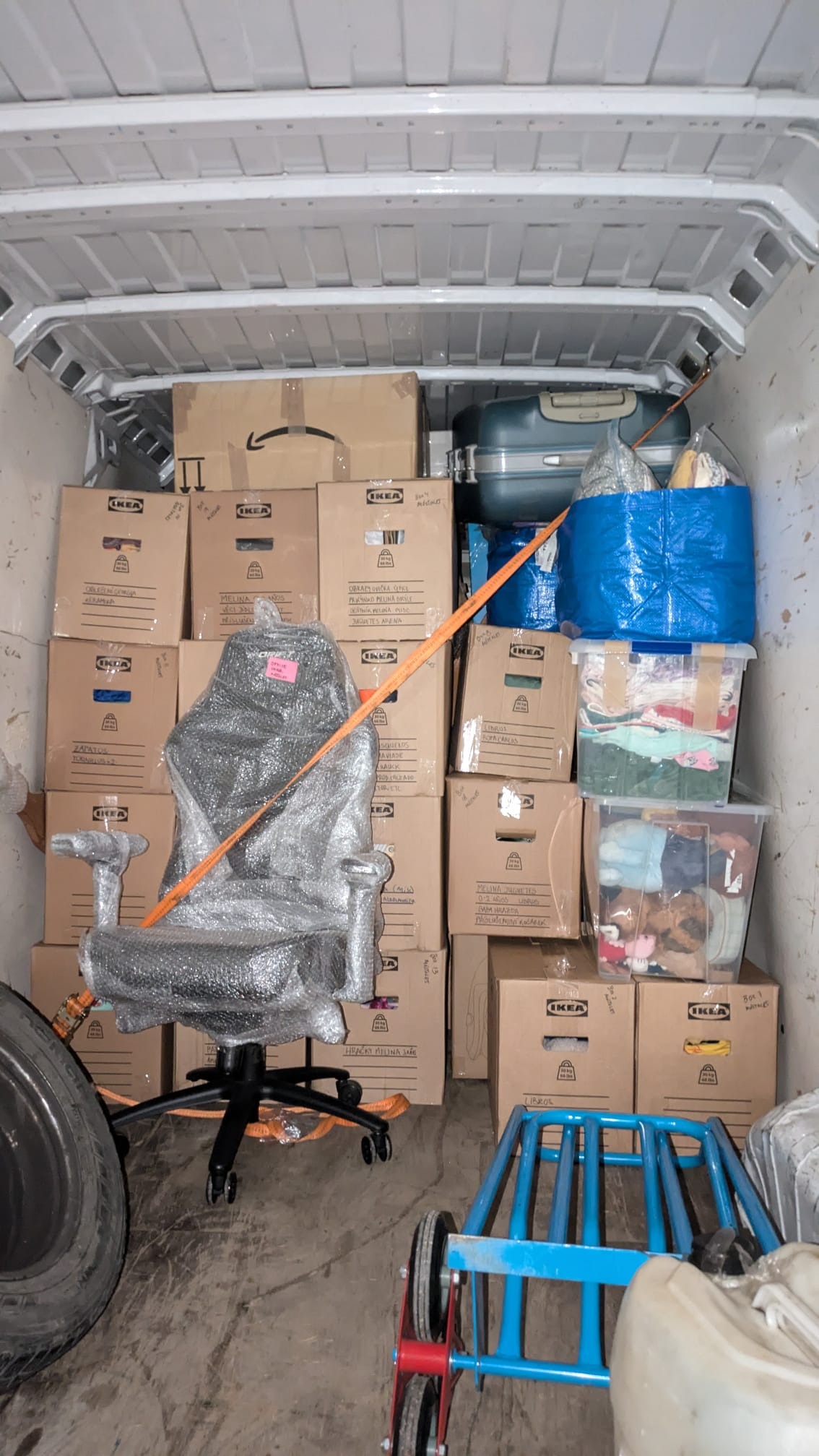 Wnętrze busa wypełnione kartonami IKEA, zabezpieczonym fotelem biurowym w folii i innymi przedmiotami, spięte pomarańczowym pasem transportowym. Przygotowanie do transportu.