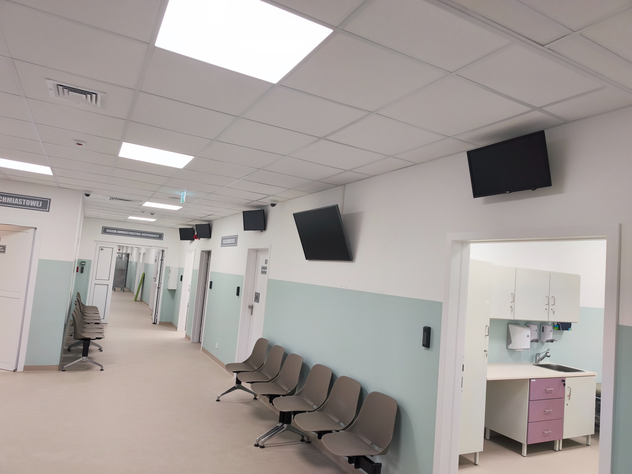 Korytarz w placówce medycznej z zamontowanymi monitorami na ścianach, rzędem krzeseł dla pacjentów i umywalką w tle. Widoczny system kontroli dostępu.