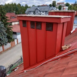 DamiXdach - Czerwony komin wentylacyjny z blachy z trzema prostokątnymi kratkami, precyzyjnie wkomponowany w dach pokryty czerwoną dachówką ceramiczną. Widoczna lina zabezpieczająca i fragmenty zabudowy...