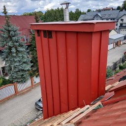 DamiXdach - Czerwona, pionowa obudowa komina z blachy, z metalowym zwieńczeniem, widoczna z góry na tle częściowo pokrytego czerwoną dachówką dachu z drewnianymi łatami, w otoczeniu domów jednorodzinnych.
