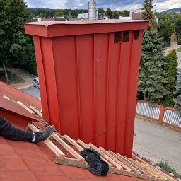 DamiXdach - Duży, czerwony komin z blachy trapezowej na dachu pokrytym czerwoną dachówką, z metalowym zwieńczeniem i widoczną nogą pracownika na drewnianej konstrukcji.