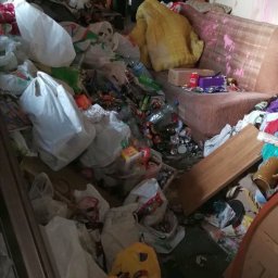 Fast Emptying - Zaniedbane wnętrze pokoju z sofą, zasypane stertą śmieci, plastikowych toreb, butelek i puszek. Widoczne elementy wyposażenia w nieładzie. Brak dbałości o porządek.