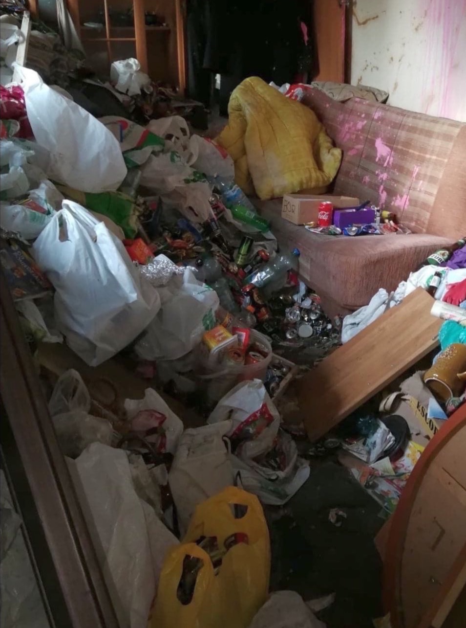Zaniedbane wnętrze pokoju z sofą, zasypane stertą śmieci, plastikowych toreb, butelek i puszek. Widoczne elementy wyposażenia w nieładzie. Brak dbałości o porządek.