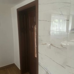 Wykończone wnętrze: drewniane drzwi z szybką i ściana z białych płytek imitujących marmur z brązowymi żyłami. Widoczny fragment podłogi z paneli.