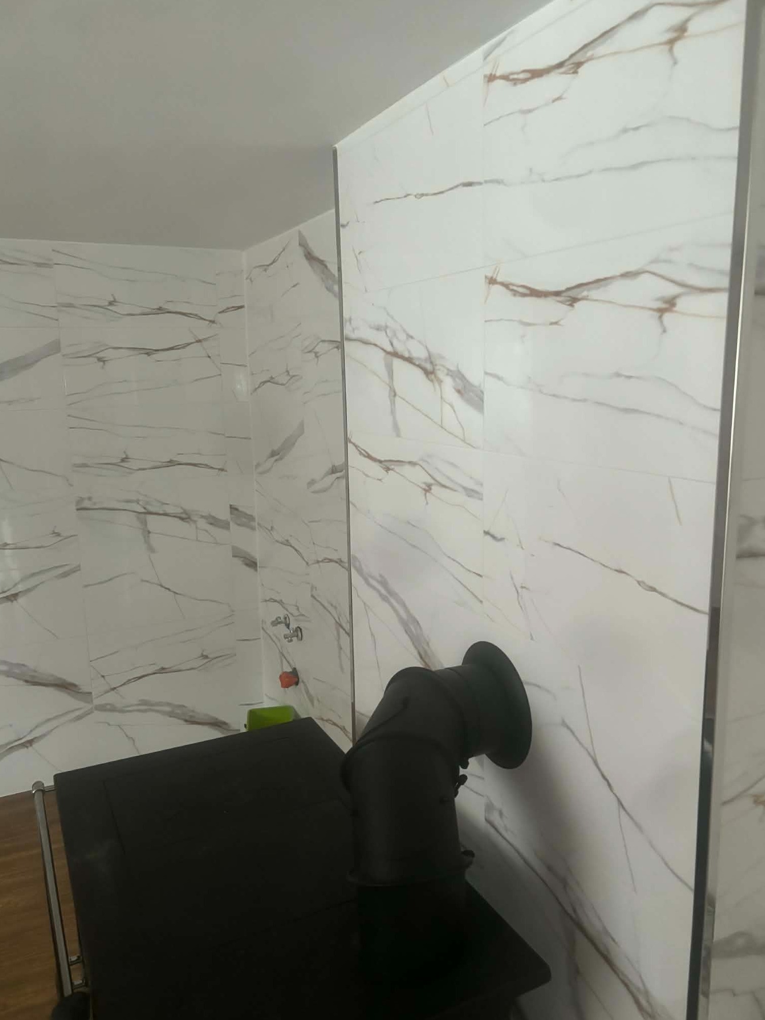 Wykończona łazienka z białymi płytkami imitującymi marmur, czarny piec typu koza z rurą odprowadzającą dym. Widoczne drewniane elementy podłogi.