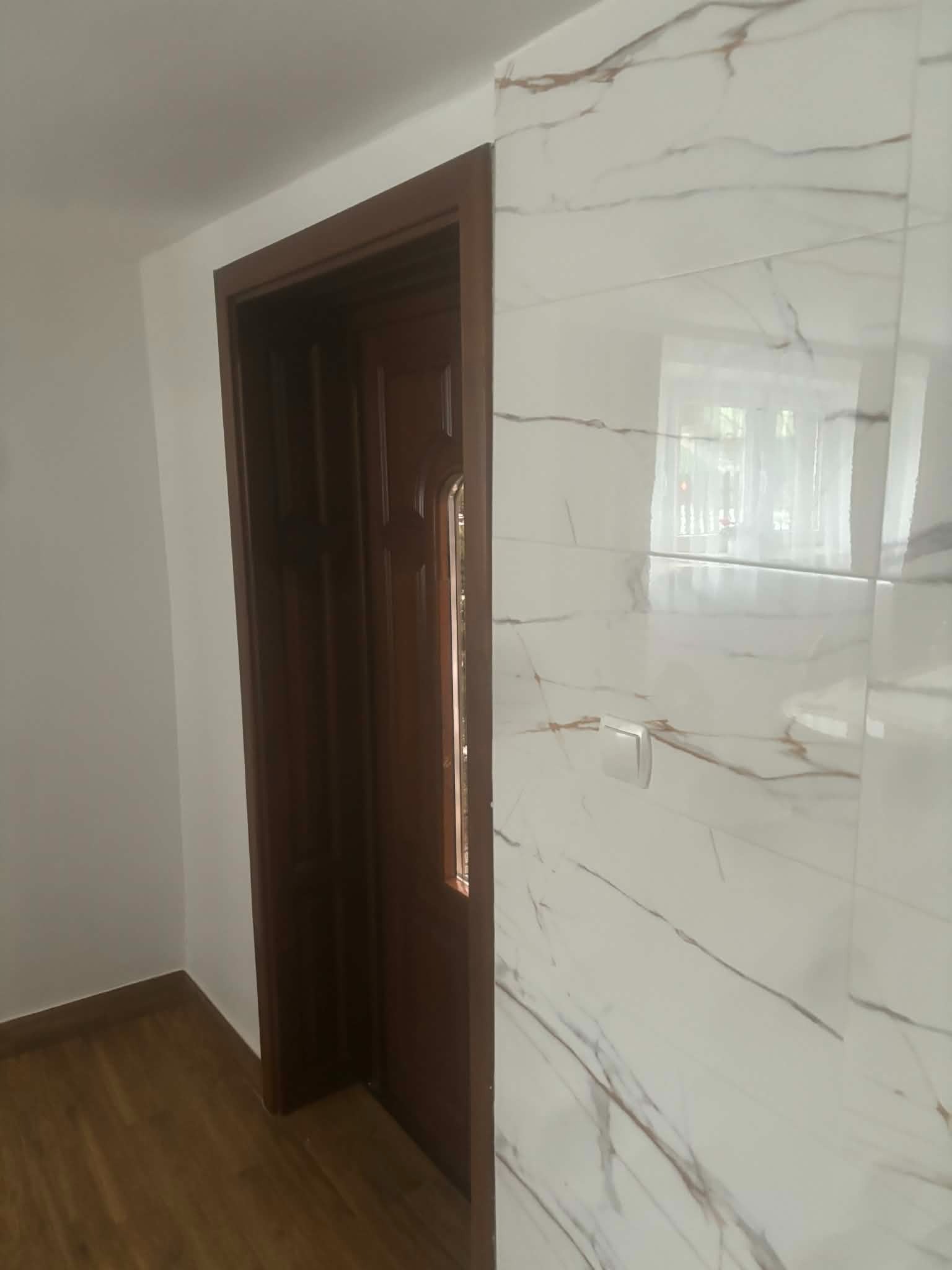 Wykończone wnętrze: drewniane drzwi z szybką i ściana z białych płytek imitujących marmur z brązowymi żyłami. Widoczny fragment podłogi z paneli.