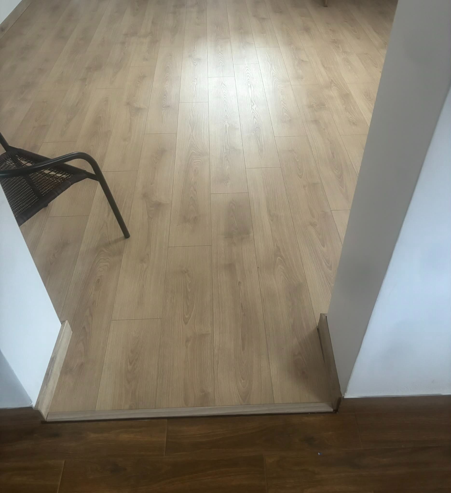 Jasne panele podłogowe ułożone wzdłuż, widoczne z perspektywy wejścia do pokoju, z fragmentem krzesła z rattanu po lewej stronie. Listwa przypodłogowa w kolorze paneli.