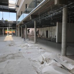 MM-Invest - z pasją i pod klucz - Wnętrze w trakcie prac wykończeniowych: widoczne kolumny, ruszt pod sufitem, zabezpieczona podłoga, drabiny i pracownicy na wysokościach. Duża przestrzeń z oknami.