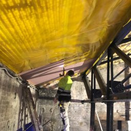 MM-Invest - z pasją i pod klucz - Pracownik montuje izolację pod żółtą folią na poddaszu w Lesznie. Widoczna drabina i konstrukcja dachu. Ceglane ściany w tle.