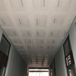 MM-Invest - z pasją i pod klucz - Sufit wykończony płytami Aquapanel w korytarzu budynku. Widoczne łączenia płyt i fragment instalacji elektrycznej. W tle okna i sylwetka osoby w odblaskowej kamizelce.
