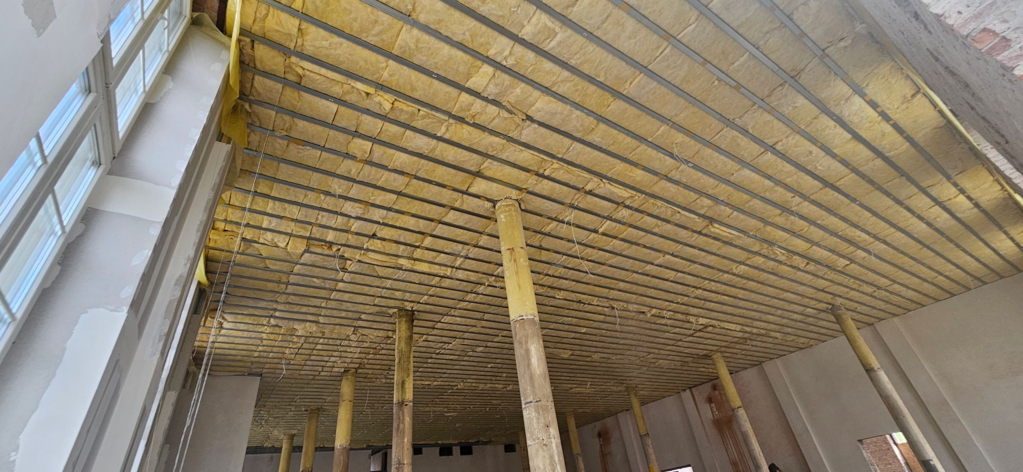 Sufit podwieszany z izolacją z wełny mineralnej, widok z dołu, metalowa konstrukcja, podpory tymczasowe, surowy stan wykończenia, okno z lewej strony.