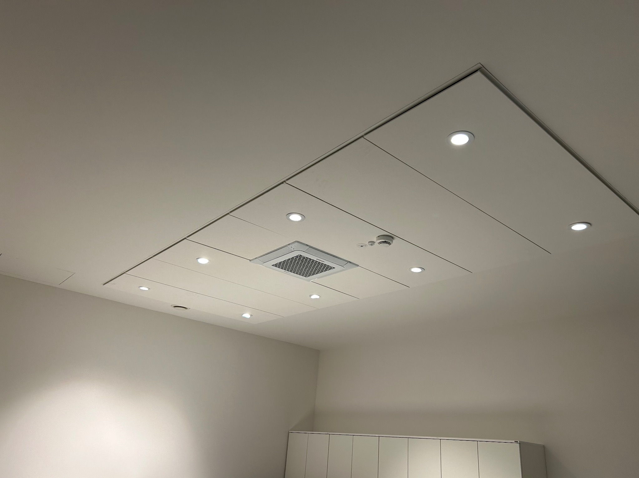 Biały sufit podwieszany z wbudowanym oświetleniem LED i kratką wentylacyjną, widok z dołu, minimalistyczny design, jasne wnętrze.