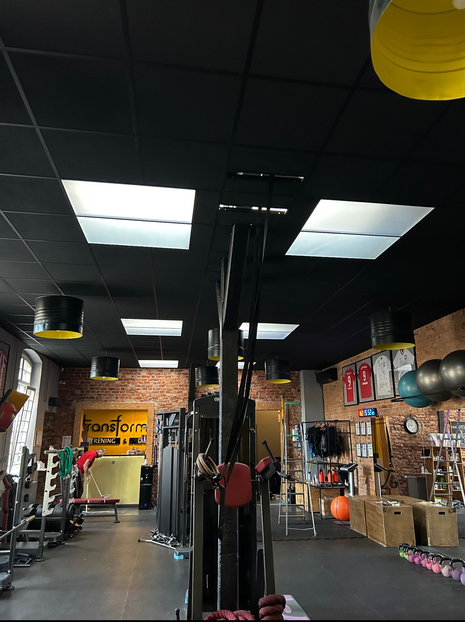 Wnętrze siłowni z czarnym sufitem, oświetleniem w stylu industrialnym i ceglanymi ścianami. Widoczne sprzęty do ćwiczeń i elementy dekoracyjne, takie jak koszulki sportowe w ramkach.