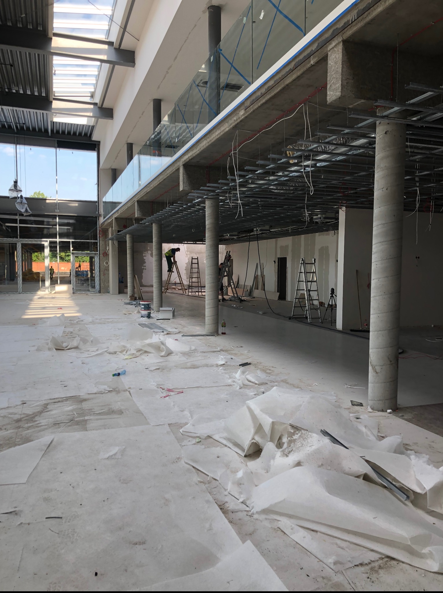 Wnętrze w trakcie prac wykończeniowych: widoczne kolumny, ruszt pod sufitem, zabezpieczona podłoga, drabiny i pracownicy na wysokościach. Duża przestrzeń z oknami.