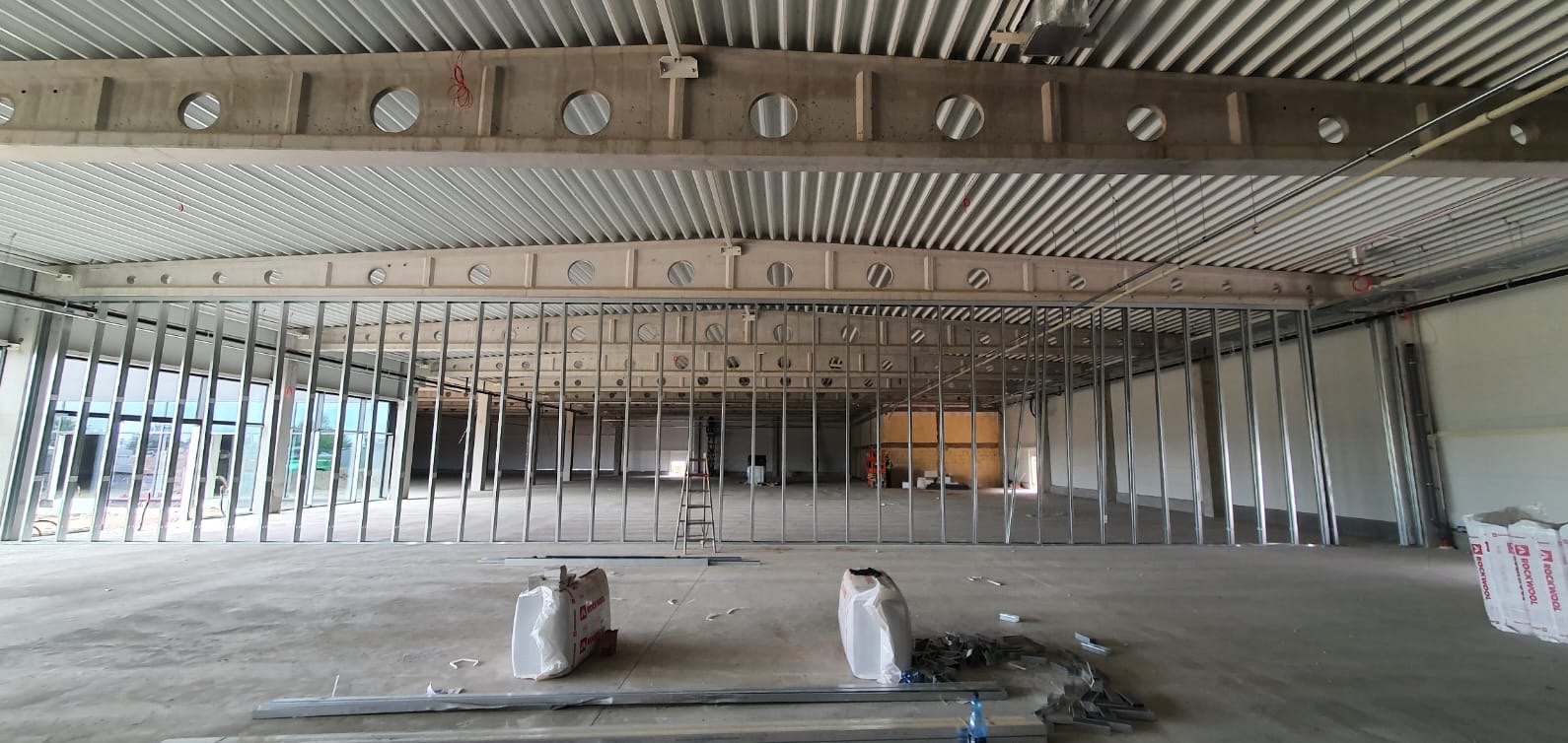 Montaż metalowej konstrukcji ścian działowych w przestronnym, surowym wnętrzu. Widoczne worki z materiałem izolacyjnym i drabina. Oświetlenie górne, betonowe elementy konstrukcyjne.