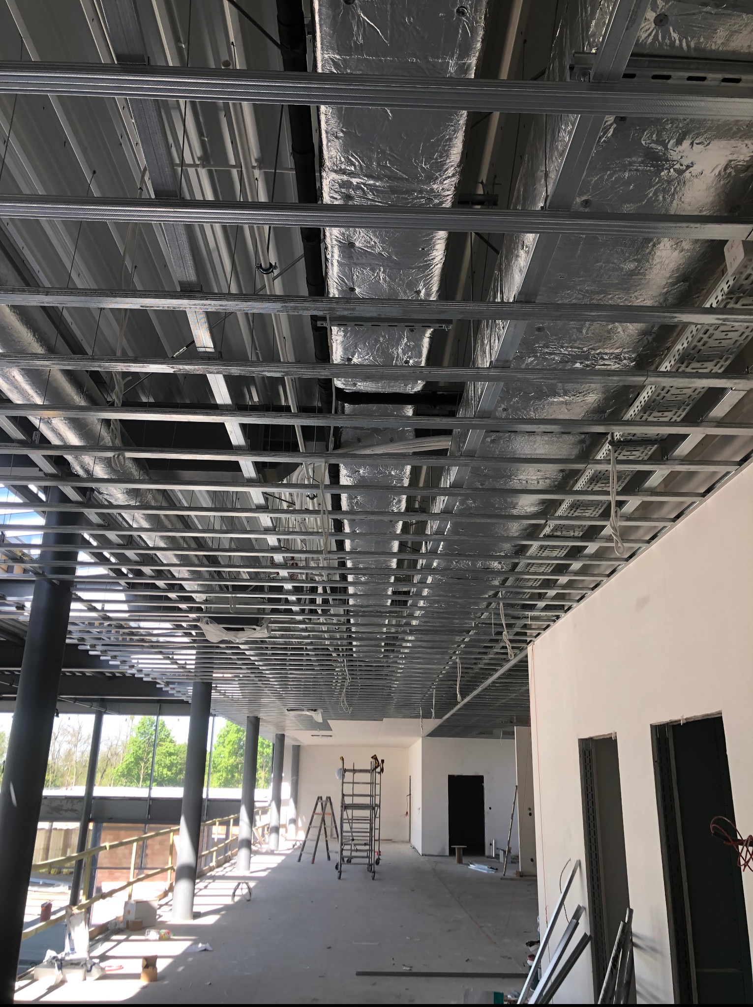 Metalowa konstrukcja sufitu podwieszanego w trakcie montażu, widoczne przewody wentylacyjne i instalacje. W tle jasne ściany i okna z widokiem na zewnątrz.
