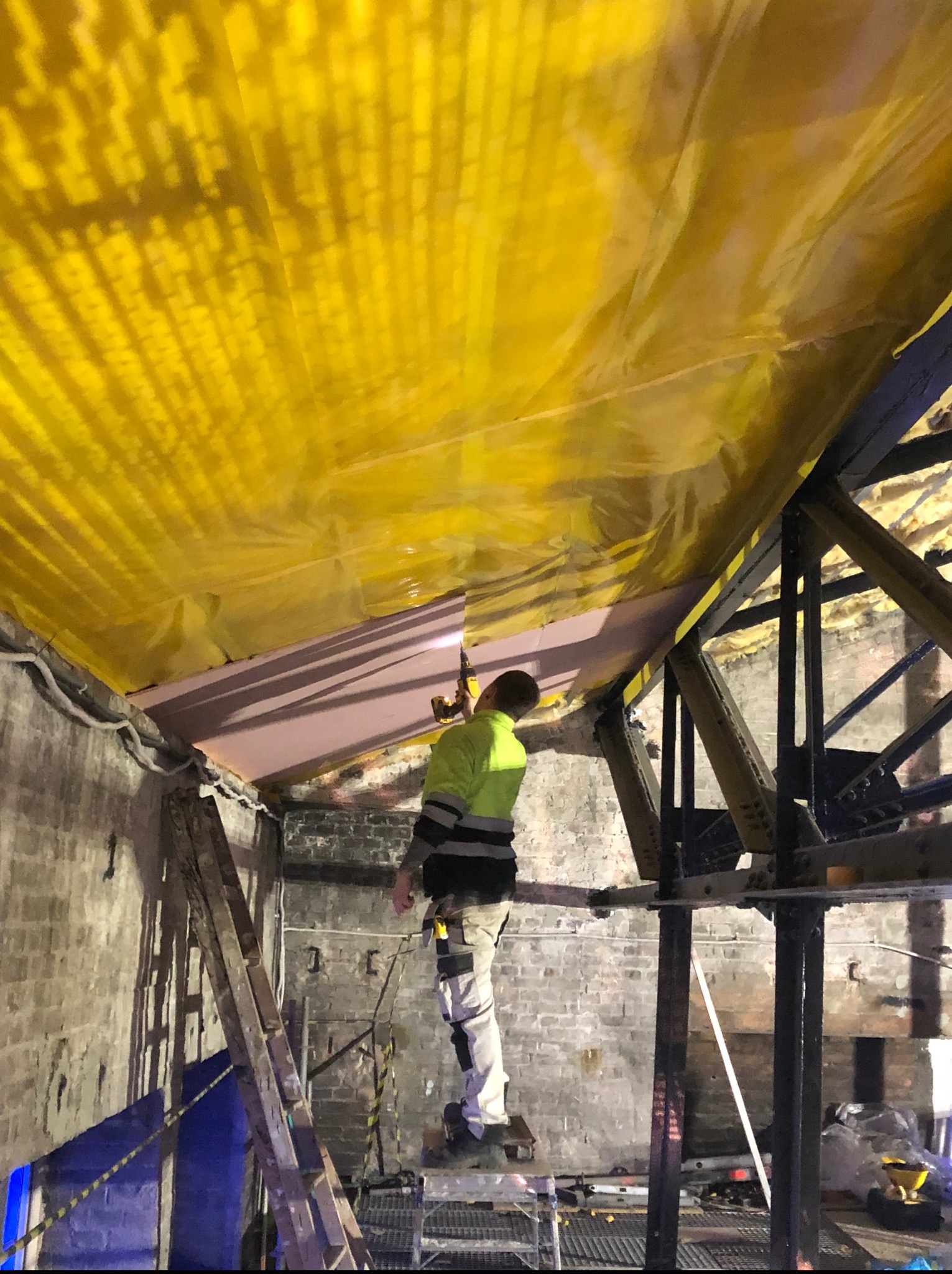 Pracownik montuje izolację pod żółtą folią na poddaszu w Lesznie. Widoczna drabina i konstrukcja dachu. Ceglane ściany w tle.