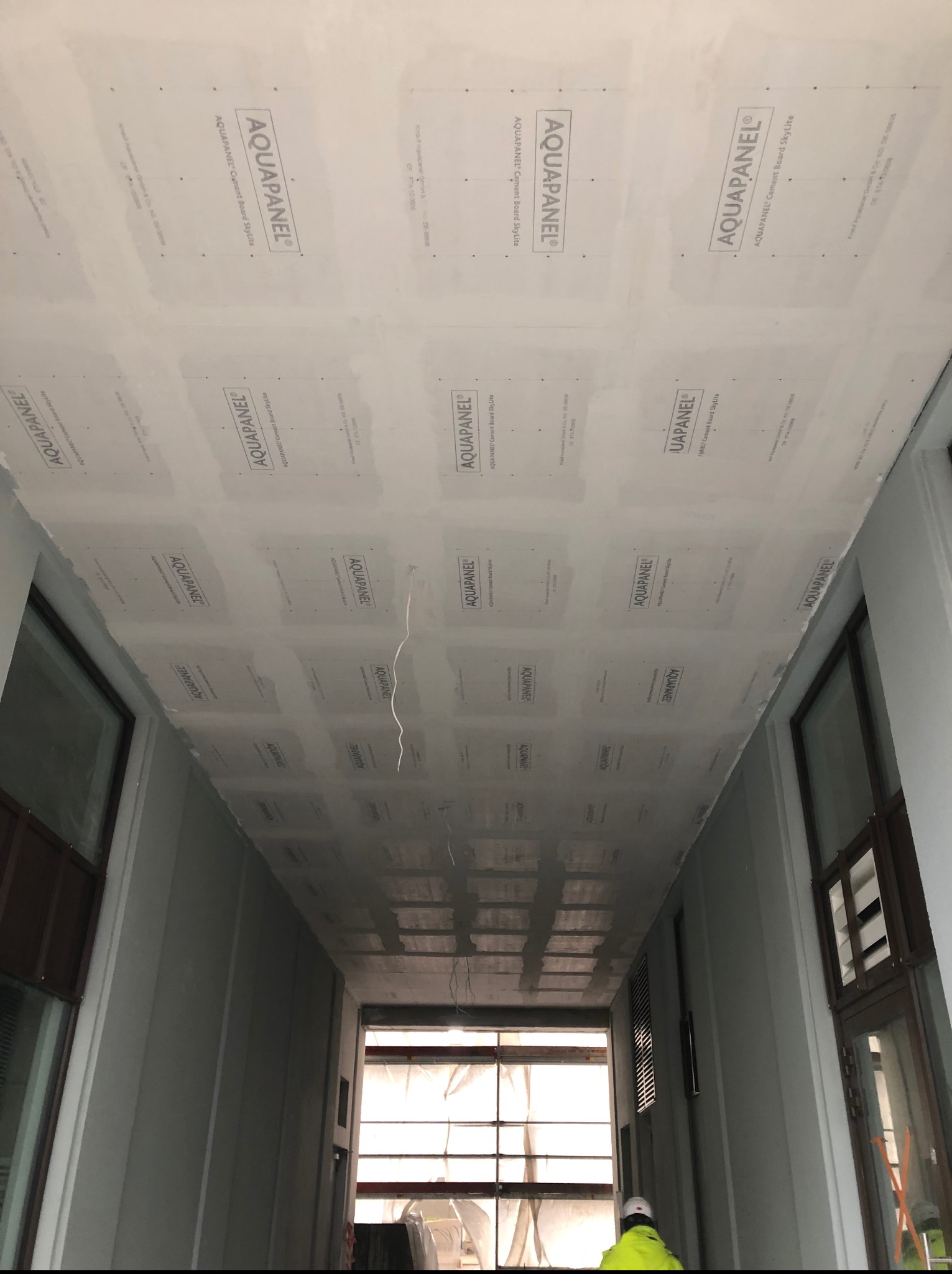Sufit wykończony płytami Aquapanel w korytarzu budynku. Widoczne łączenia płyt i fragment instalacji elektrycznej. W tle okna i sylwetka osoby w odblaskowej kamizelce.