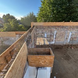 Posadzki Maszynowe - Przygotowanie szalunku i zbrojenia pod wylewkę stropu. Widoczne deski szalunkowe, stalowe pręty zbrojeniowe, rusztowanie i fragment ściany z cegły. Prace budowlane w toku.