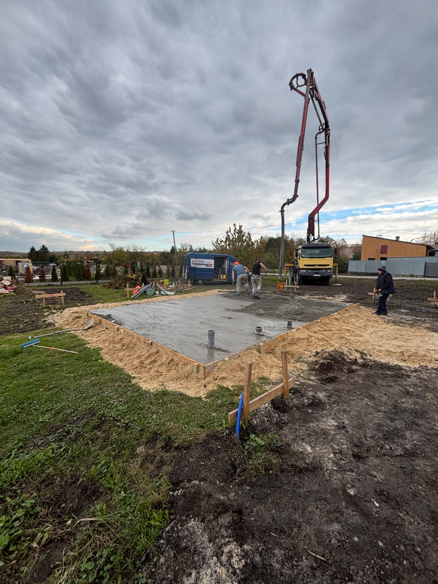 Wylewanie betonu na przygotowany fundament z szalunkiem, widoczny samochód z pompą do betonu i pracownicy w trakcie pracy, pochmurne niebo w tle.