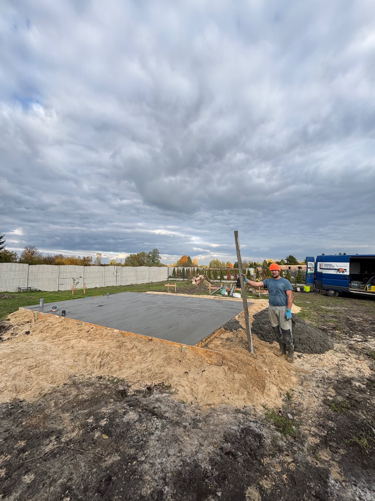 Wyrównana, szara powierzchnia w drewnianej ramie, prawdopodobnie podłoże pod budynek. Obok stoi mężczyzna z łatą murarską, w tle widoczny samochód dostawczy.