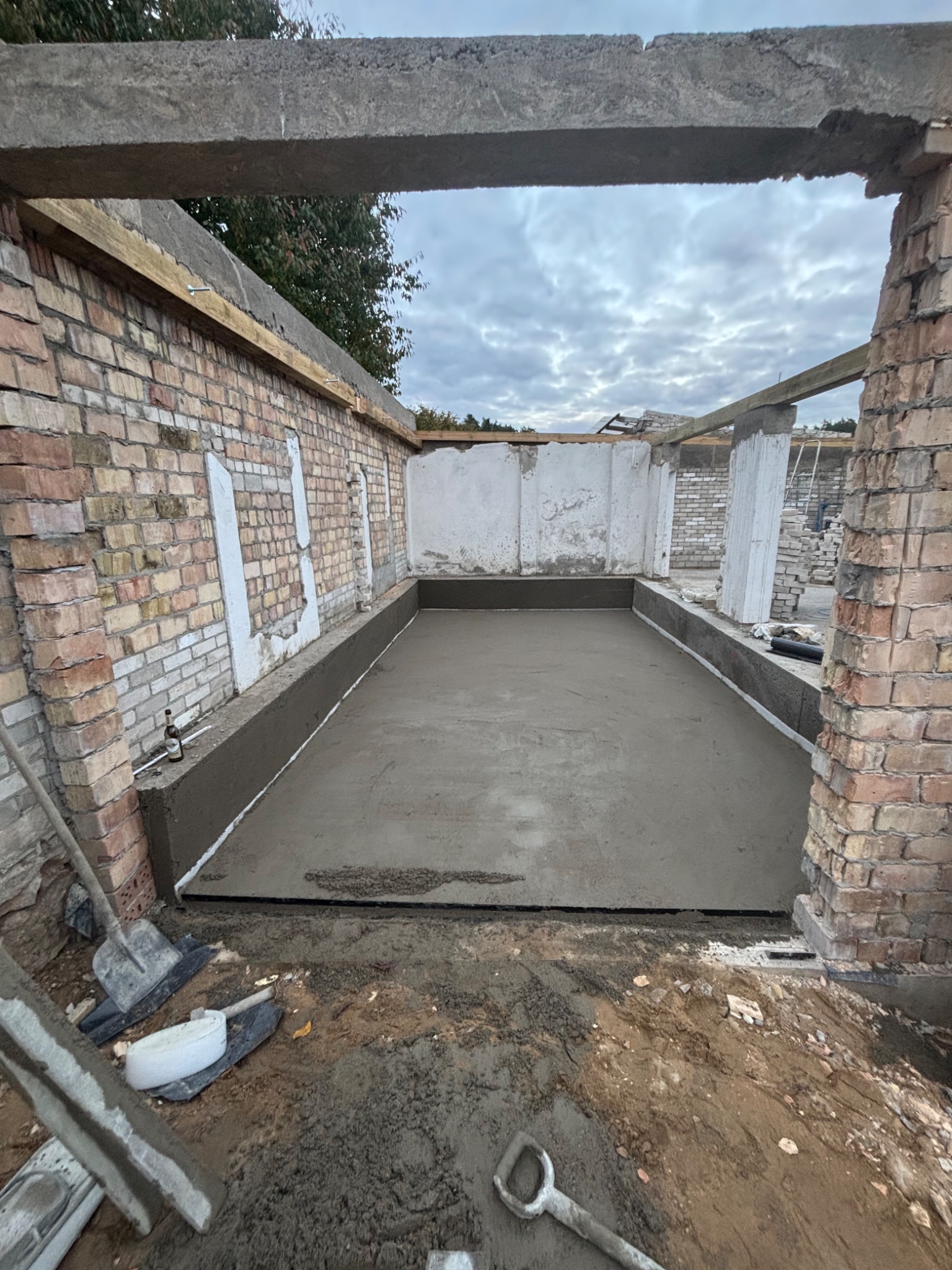 Świeżo wylana, gładka podłoga w pomieszczeniu z ceglanymi ścianami i surowym betonowym nadprożem. Widoczne narzędzia i butelka. Wnętrze w trakcie prac budowlanych.