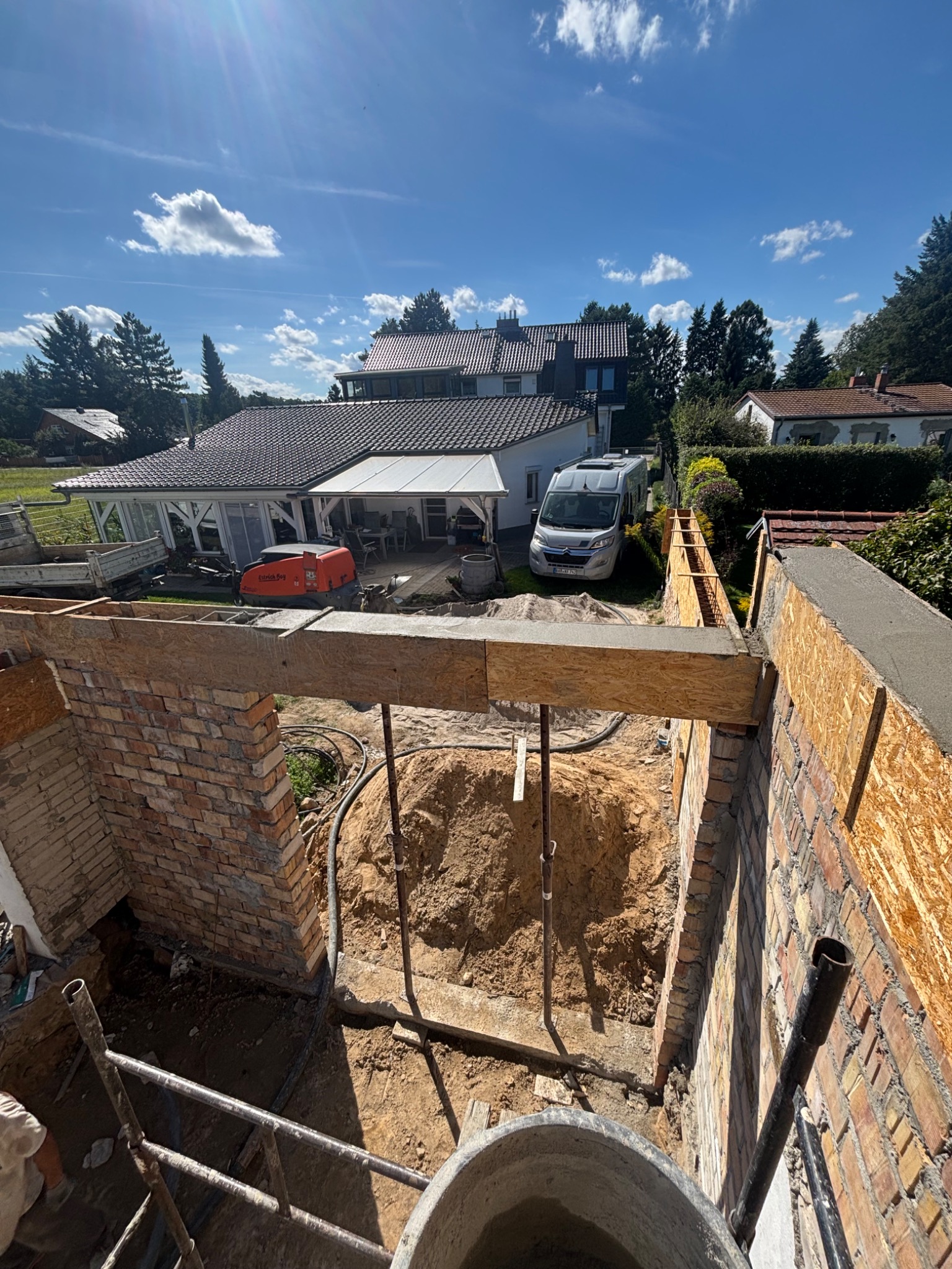 Przygotowanie podłoża pod wylewkę: widok z góry na wykop z ceglanymi ścianami, szalunek z desek i piasek. W tle domy i błękitne niebo.