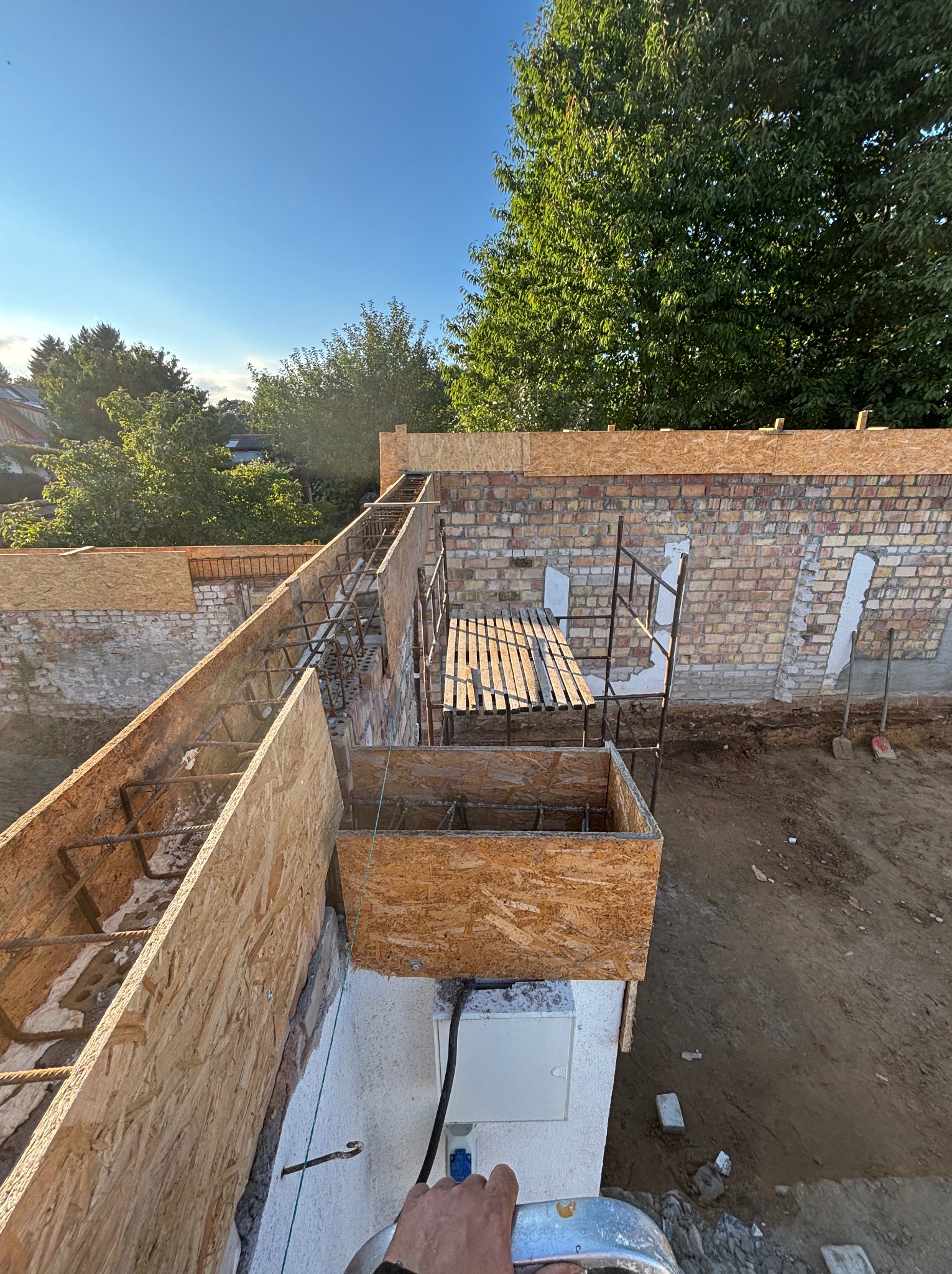 Przygotowanie szalunku i zbrojenia pod wylewkę stropu. Widoczne deski szalunkowe, stalowe pręty zbrojeniowe, rusztowanie i fragment ściany z cegły. Prace budowlane w toku.