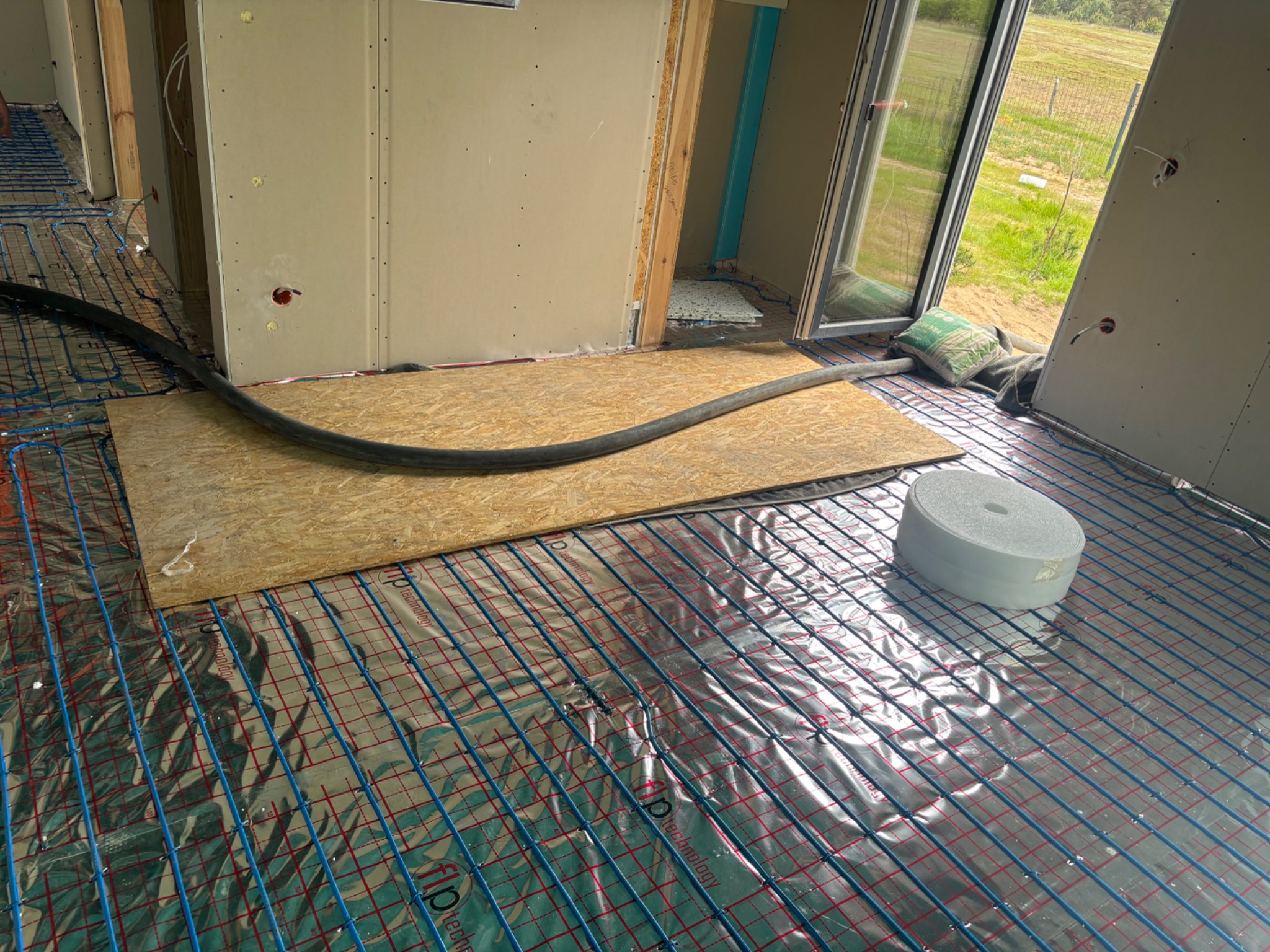 Rozłożona instalacja ogrzewania podłogowego na folii aluminiowej, z widoczną rurą doprowadzającą i płytą OSB. Przygotowanie do wylania podkładu.