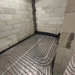 Ułożone rury ogrzewania podłogowego na folii izolacyjnej w surowym pomieszczeniu z betonowych bloczków. Widok z góry, instalacja przed wylaniem posadzki.
