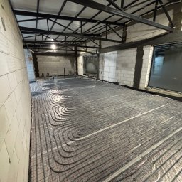 Szare rury ogrzewania podłogowego ułożone na folii z czerwoną siatką w surowym wnętrzu z białych bloczków i metalową konstrukcją sufitu. Widok z góry, szeroki kąt.