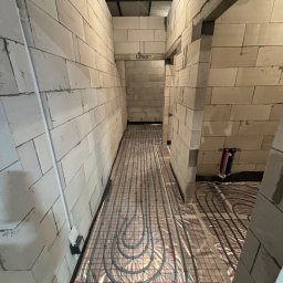 Montaż ogrzewania podłogowego w surowym wnętrzu. Szare rury ułożone na folii z czerwoną kratką, ściany z białych bloczków, widoczne instalacje hydrauliczne.