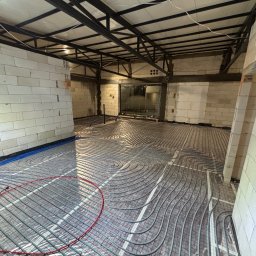 Szare rury ogrzewania podłogowego ułożone spiralnie na folii w surowym wnętrzu z białych bloczków i metalową konstrukcją sufitu. Widoczne niebieskie rury przy ścianie.